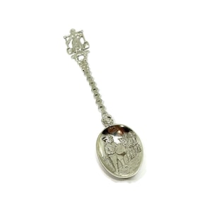 May include: A silver-colored souvenir spoon with a detailed design. The spoon features a decorative handle with a figure at the top and a chain-like design. The bowl of the spoon has an engraved scene of people. A collectible item.