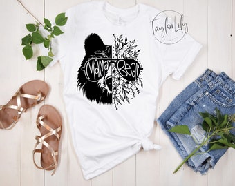 Puede incluir: Camiseta blanca con un gráfico en blanco y negro de un oso con gafas de sol y detalles florales. El texto "Mama Bear" está en el gráfico.
