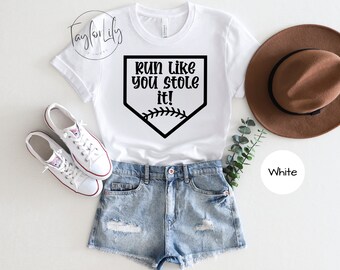 Op de afbeelding: Wit T-shirt met een baseball-diamantgrafiek en de tekst "Run like you stole it!" in zwart gedrukt. Het shirt wordt getoond op een model dat een spijkerbroek en witte sneakers draagt.