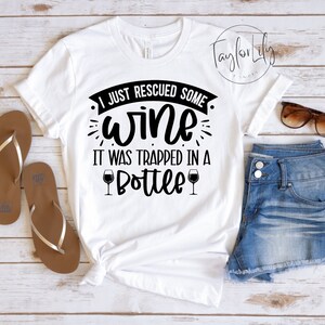 Puede incluir: Camiseta blanca con texto negro que dice "Acabo de rescatar algo de vino. Estaba atrapado en una botella." El texto está rodeado de elementos decorativos. Dos iconos de copas de vino están a cada lado de la palabra "botella".