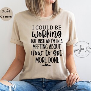 May include: A beige t-shirt with black text that reads "I could be working but instead I'm in a meeting about how to get more done".