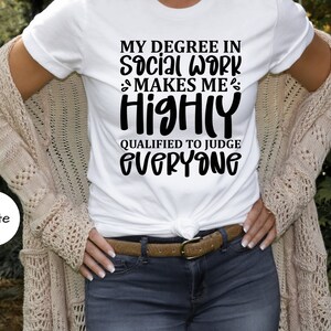 以下が含まれることがあります： 「MY DEGREE IN SOCIAL WORK MAKES ME HIGHLY QUALIFIED TO JUDGE EVERYONE」の黒い文字がプリントされた白いTシャツ。青いジーンズ、茶色のベルト、ベージュのカーディガンとコーディネートされています。