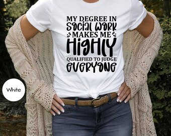 May include: White t-shirt with the text "MY DEGREE IN SOCIAL WORK MAKES ME HIGHLY QUALIFIED TO JUDGE EVERYONE" in black. The shirt is paired with blue jeans, a brown belt, and a beige cardigan.