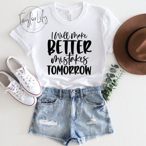 Puede incluir: Camiseta blanca con texto negro que dice "Haré mejores errores mañana". La camiseta está sobre una superficie blanca con un par de zapatillas blancas, un sombrero marrón y una rama de vegetación.