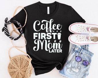 Puede incluir: Camiseta negra con texto blanco que dice "Coffee First Mom Later". La camiseta está colocada sobre una superficie blanca con un bolso de paja, unas gafas de sol y unas zapatillas blancas.