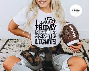 Op de afbeelding: Wit T-shirt met zwarte tekst die "I spend FRIDAY NIGHTS under the LIGHTS" zegt met een klein rood hartje en een voetbalafbeelding. Het model houdt een bruin-witte voetbal vast.