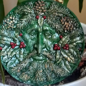 May include: A green, ceramic, round ornament with a detailed, sculpted face of the Green Man. The face is surrounded by leaves, berries, and pine cones.