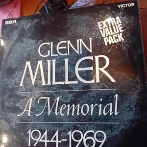 Pode incluir: Um álbum de vinil vintage de Glenn Miller intitulado "A Memorial" com os anos 1944-1969. A capa do álbum é preta com letras brancas e uma aparência texturizada e manchada. Os logotipos da RCA e Victor são visíveis.