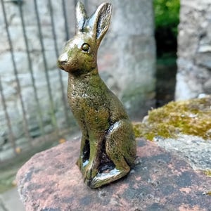 Puede incluir: Una figurita de conejo de color bronce con una superficie detallada y texturizada. El conejo está sentado en posición vertical con las orejas levantadas.