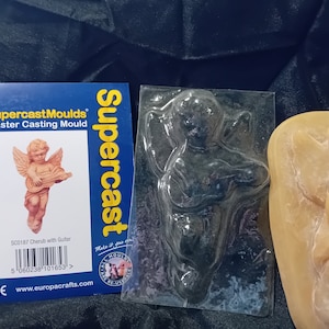 Puede incluir: Un molde de plástico transparente de un querubín tocando la guitarra. El molde está al lado de una fundición de yeso del querubín. El embalaje del molde es visible en el fondo. El embalaje es amarillo y negro y dice "Supercast Moulds" y "Molde de fundición de yeso".