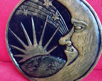 Pode incluir: Uma decoração de parede circular, cor de bronze, com um design celestial. A obra de arte retrata uma lua crescente com um rosto, estrelas e um sol radiante contra um fundo escuro. Uma corda verde está presa para pendurar.