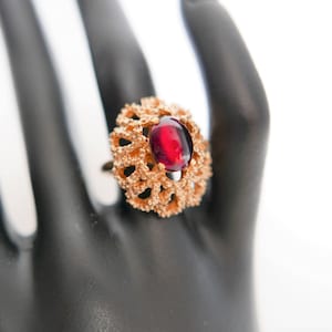 Op de afbeelding: Een gouden ring met een rode edelsteen gezet in een delicaat, ingewikkeld ontwerp. De ring is op een zwarte displayhand.