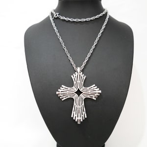 Op de afbeelding: Een zilverkleurige hangerketting met een groot, gestileerd kruisontwerp. Het kruis heeft een gelaagde, open constructie met gebogen en rechte lijnen. De ketting wordt getoond op een zwarte mannequin.