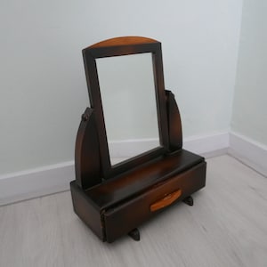 Peut inclure: Un miroir de toilette en bois brun foncé avec un petit tiroir. Le miroir a un bord biseauté et est encadré d'un design simple et élégant.