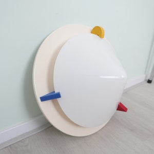 Könnte beinhalten: Eine moderne Wandleuchte mit cremefarbenem, kreisförmigem Sockel und einem weißen, abgerundeten Leuchtenschirm. Sie verfügt über geometrische Akzente in Blau, Rot und Gelb. Das Design ist minimalistisch und künstlerisch, geeignet für die Inneneinrichtung.