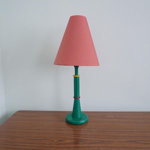 Puede incluir: Una lámpara de mesa verde con una pantalla rosa. La lámpara tiene una base cilíndrica con anillos de colores alrededor.