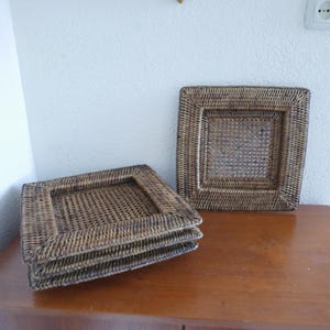 Op de afbeelding: Vier vierkante placemats van geweven rotan in verschillende bruintinten. De placemats hebben een vierkante vorm met een geweven midden en een dikkere rand. De placemats zijn gestapeld op een houten oppervlak.