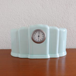 Puede incluir: Un reloj de cerámica azul claro con una esfera redonda y agujas negras. El reloj es de estilo art déco con una forma rectangular redondeada.