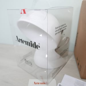Puede incluir: Una lámpara de mesa blanca con una pantalla redonda en forma de hongo. La lámpara está empaquetada en una caja de plástico transparente con el nombre de la marca "Artemide" impreso en el lateral. La caja también incluye el texto "Quasi un cappuccino con un foglio stampato per una luce perfetta."