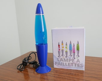May include: A blue lava lamp with a silver base and a cord. The lamp is on a wooden surface. The box behind the lamp is white with the text "Lampe a Paillettes" in black.