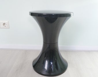 Könnte beinhalten: Ein schwarzer Plastikhocker mit einer breiten, runden Oberseite und einem ausgestellten Fuß. Der Hocker ist aus glattem, glänzendem Kunststoff.