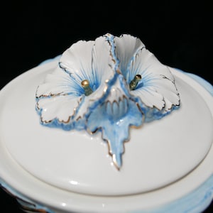 May include: White porcelain lid with a blue and white floral design. The flower is three-dimensional and has gold accents.