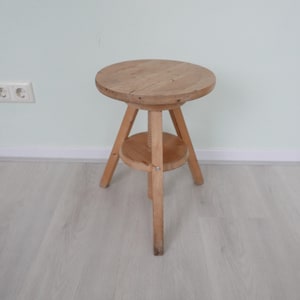 Könnte beinhalten: Ein hellfarbiger Holzstuhl mit rundem Sitz und drei Beinen. Der Hocker hat ein unteres Regal und eine zentrale Gewindeunterstützung. Das Holz hat sichtbare Maserung und Knoten, was ihm ein rustikales Aussehen verleiht. Der Hocker ist etwa 46 cm hoch.