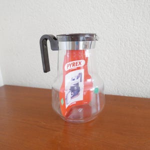 Peut inclure: Une carafe en verre transparent avec une poignée en plastique marron et un couvercle argenté. La carafe porte une étiquette "PYREX".