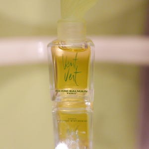 Puede incluir: Un pequeño frasco de perfume transparente con un líquido amarillo-verdoso. El frasco tiene una tapa verde claro y las palabras "Vent Vert" en escritura verde. La marca "PIERRE BALMAIN PARIS" está impresa debajo.