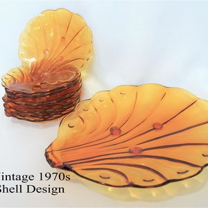 Op de afbeelding: Amberkleurige vintage glazen borden met een schelpontwerp. De borden hebben een geschulpte rand en een waaiervormig patroon, met kleine, ronde accenten. De tekst "Vintage 1970s Shell Design" is zichtbaar.