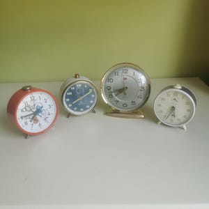 Könnte beinhalten: Vier Vintage-Wecker in verschiedenen Größen und Designs. Die Uhren haben runde Zifferblätter mit Zahlen und Zeigern. Einer ist rot mit einer Zeichentrickfigur, ein anderer hat ein blaues Zifferblatt, und eine goldfarbene Uhr sticht hervor.