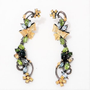 Op de afbeelding: Een paar oorhangers met een gouden strik en groene, blauwe en zwarte edelstenen. De oorhangers zijn gemaakt van zilver en hebben een bloemmotief.