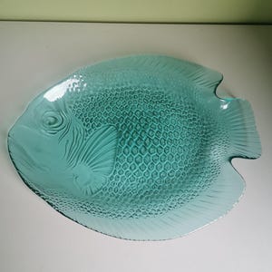 Op de afbeelding: Een turquoise glazen serveerschaal in de vorm van een vis. De schaal heeft gedetailleerde schubben en vinnen, met een textuur. Deze visvormige schaal is een decoratief en functioneel item.