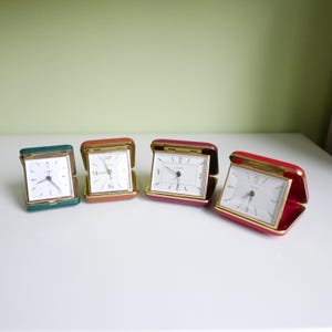 Puede incluir: Cuatro relojes despertadores vintage de diferentes colores: verde, marrón, rosa y rojo. Cada reloj tiene una esfera blanca con números y manecillas negras.