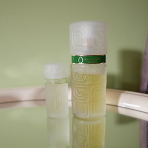 Op de afbeelding: Twee parfumflacons van matglas met witte deksels. De grotere fles heeft een groene band met de tekst "DE LANCOME". De flessen hebben een vierkant patroon. Het parfum in de flessen is geel.