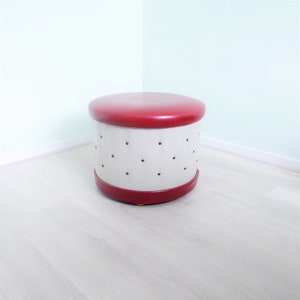 May include: A white and red round storage ottoman with a red top and a white base with small holes. The ottoman is sitting on a light wood floor.