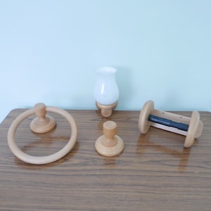 May include: A set of light-coloured wooden bathroom accessories. The collection includes a towel ring, a small hook, a wall-mounted light fixture with a white glass shade, and a toilet paper holder. The items are displayed on a wooden surface.