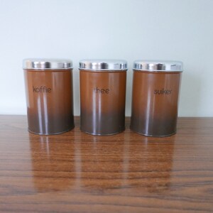 Op de afbeelding: Drie bruine metalen kannen met zilveren deksels. De kannen zijn gelabeld met "koffie", "thee" en "suiker".