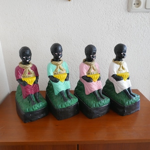 Könnte beinhalten: Vier schwarze Figuren von Kindern, die auf grünen Sockeln sitzen, jeweils eine gelbe Schale halten und ein andersfarbiges Gewand tragen: Burgunderrot, Türkis, Rosa und Weiß. Jede Figur trägt das Wort "MERCI" auf ihrem Gewand.