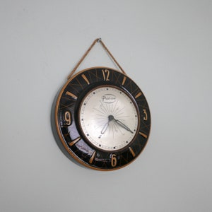 May include: A black and gold round clock with Roman numerals and a white face. The clock has a black and gold design with a thin rope hanging from the top.