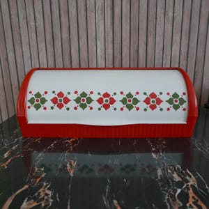 Op de afbeelding: Een rode en witte retro broodtrommel met een bloemenpatroon. De rechthoekige container heeft afgeronde uiteinden en een geribbelde basis. Het witte gedeelte toont een herhalend ontwerp van rode en groene bloemen en stippen.