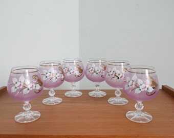 May include: Set of six pink glass goblets with gold trim and white floral designs. The goblets are arranged in a row on a wooden surface.