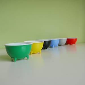 May include: A series of seven small, brightly coloured bowls with white interiors and tripod bases. The bowls are in a range of colours: green, yellow, black, blue, grey, red, and white. They are arranged in a row on a white surface.