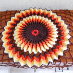 Può includere: Un centrino all'uncinetto con un centro marrone e un design multicolore di arancione, giallo e bianco. Il centrino è appoggiato su una superficie a rete marrone.