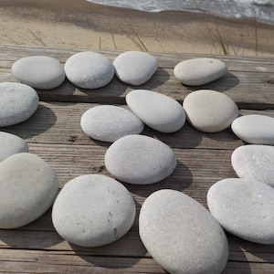May include: A collection of smooth, gray river stones arranged on a weathered wooden surface. The stones vary in size and shape, with a natural, organic aesthetic. The background shows a beach and ocean.