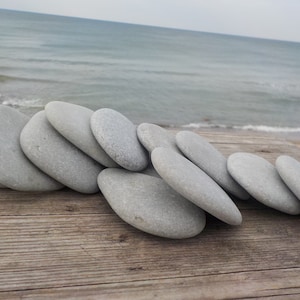 May include: A collection of smooth, gray stones arranged on a weathered wooden surface. The stones are oval-shaped and vary slightly in size. The background features a calm ocean and a cloudy sky, creating a natural, serene scene.