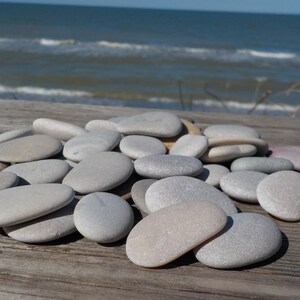 May include: A pile of smooth, gray and white stones, likely collected from a beach, arranged on a wooden surface. The stones are oval-shaped and have a natural, polished look.