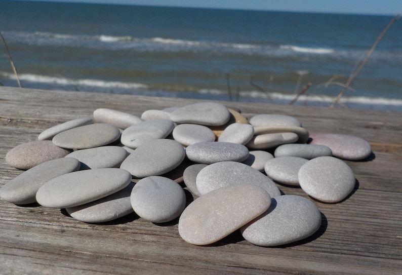 May include: A pile of smooth, gray and white stones, likely collected from a beach, arranged on a wooden surface. The stones are oval-shaped and have a natural, polished look.