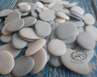 May include: A pile of smooth, gray and white stones, some with a slightly rough texture. The stones are oval and round in shape, and they are arranged in a random pattern. The stones are on a blue wooden surface.