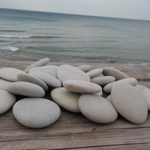 May include: A pile of smooth, light gray stones on a weathered wooden surface. The stones are various sizes and shapes, with a slightly rounded oval form. The background is a blurry image of a beach and ocean.